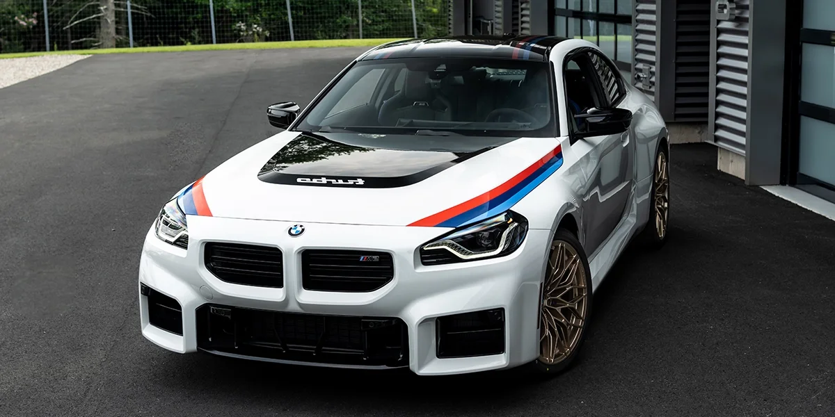2026 BMW M2 Turbo Coupe in Alpine White with M Performance stripes and gold wheels parked outside BMW facility.