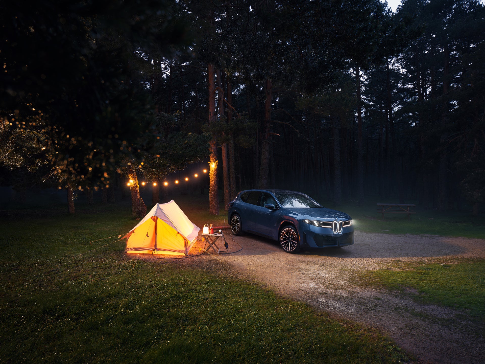 BMW iX3 beside an illuminated tent at a wooded campsite