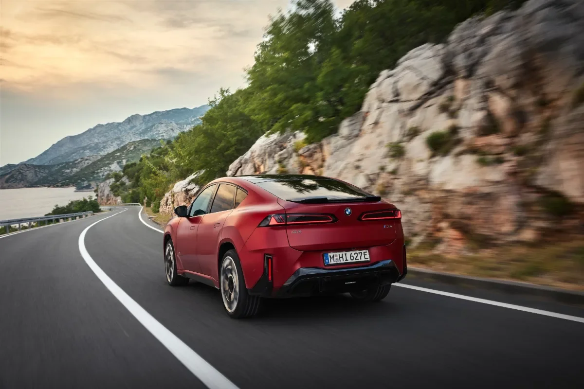 Red BMW iX4 M60 xDrive driving on a winding coastal mountain road.